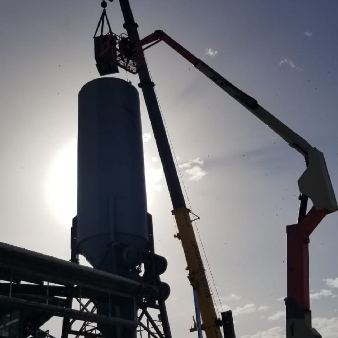 Cylindrical steel silo being lifted by crane against the sun